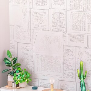 May include: A white dresser with two drawers, a small potted plant, a larger potted plant, a small blue speaker, a lamp with a clear glass bulb, and a woven placemat. The wall behind the dresser is covered in a white wallpaper with a pattern of faded newspaper text.