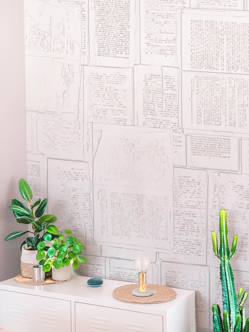 May include: A white dresser with two drawers, a small potted plant, a larger potted plant, a small blue speaker, a lamp with a clear glass bulb, and a woven placemat. The wall behind the dresser is covered in a white wallpaper with a pattern of faded newspaper text.
