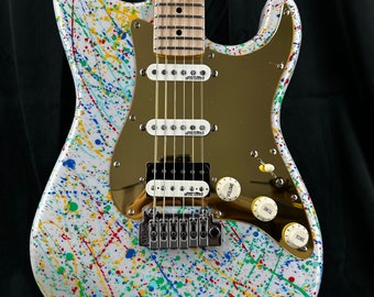Op de afbeelding: Een witte elektrische gitaar met een gouden slagplaat en een kleurrijk splatterontwerp. De gitaar heeft een esdoornhals en een palissander toets.