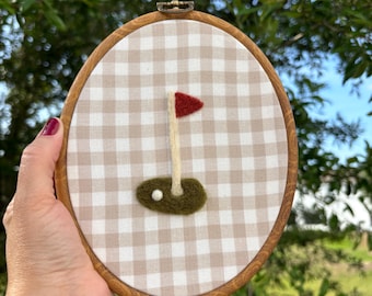 May include: An oval-shaped embroidery hoop featuring a golf-themed design. The artwork includes a red flag, a white flagpole, a green patch of grass, and a white golf ball. The background is a tan and white gingham pattern, framed by a wooden hoop.