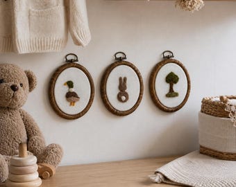 May include: Three framed embroidered artworks with wooden oval frames. The artworks feature a duck, a rabbit, and a tree on a white background. The frames are hanging on a white wall. A teddy bear and a wooden stacking toy are on a wooden surface.