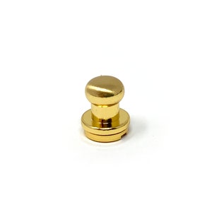 May include: A gold-colored metal button with a round top and a flat base. The button has a small hole in the center of the base.