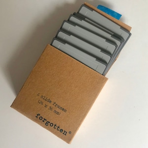 Peut inclure: Une boîte en carton marron avec une étiquette grise et blanche indiquant "6 slide frames (24 x 36 mm) forgotten®". La boîte contient six cadres à diapositives gris.