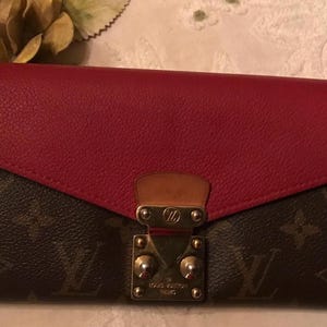 May include: A red and brown leather wallet with a gold clasp and the Louis Vuitton logo. The wallet has a flap closure and a brown monogram pattern.