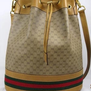 May include: Tan bucket bag with a repeating logo pattern. The bag has a tan leather drawstring closure and shoulder strap. A red and green stripe detail accents the bottom of the bag. Gold-tone hardware is visible.