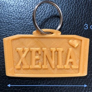 Puede incluir: Un llavero de color naranja claro con el nombre "XENIA" en letras en relieve. El llavero rectangular mide aproximadamente 6 cm de largo y 3 cm de alto, con un peque&ntilde;o detalle de coraz&oacute;n. Un llavero plateado est&aacute; adjunto en la parte superior.