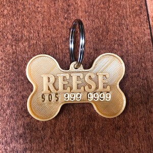 Puede incluir: Una etiqueta para mascotas en forma de hueso de color dorado claro, con el nombre "REESE" y un n&uacute;mero de tel&eacute;fono impreso. La etiqueta est&aacute; sujeta a un anillo de metal plateado.