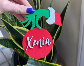 Puede incluir: Adorno de mano verde del Grinch con un gorro de Papá Noel blanco que sostiene un adorno rojo con el nombre "Xenia" escrito en blanco.