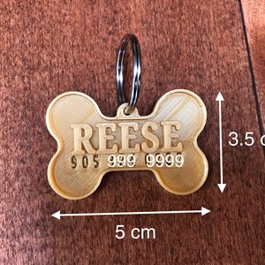 Puede incluir: Una placa para mascotas en forma de hueso, de color beige, con el nombre "REESE" y un n&uacute;mero de tel&eacute;fono. La placa mide 5 cm de largo y 3,5 cm de ancho, con un anillo de metal plateado.