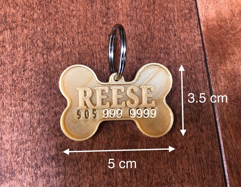 Puede incluir: Una placa para mascotas en forma de hueso, de color beige, con el nombre "REESE" y un n&uacute;mero de tel&eacute;fono. La placa mide 5 cm de largo y 3,5 cm de ancho, con un anillo de metal plateado.