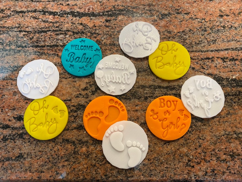 Pode incluir: Nove cortadores de biscoitos redondos com v&aacute;rios desenhos tem&aacute;ticos de beb&ecirc;. Os desenhos incluem "Bem-vindo beb&ecirc;", "Oh beb&ecirc;", "Menino ou menina?", "Voc&ecirc; &eacute; meu sol" e pegadas. Os cortadores s&atilde;o de v&aacute;rias cores, incluindo branco, amarelo, laranja e azul.