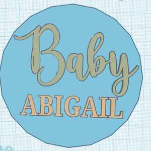 Pode incluir: Um c&iacute;rculo azul claro com o texto "Baby ABIGAIL" em bege e cinza.