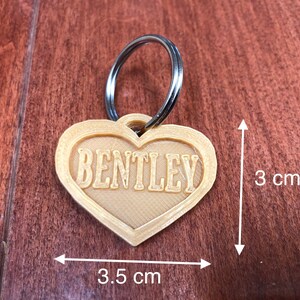 Puede incluir: Una etiqueta para mascotas en forma de coraz&oacute;n con el nombre "BENTLEY" en una fuente serif. La etiqueta mide aproximadamente 3,5 cm de ancho y 3 cm de alto, con un llavero plateado adjunto. La etiqueta es de color beige claro y est&aacute; sobre una superficie de madera.