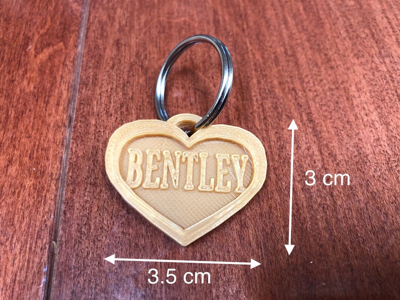 Puede incluir: Una etiqueta para mascotas en forma de coraz&oacute;n con el nombre "BENTLEY" en una fuente serif. La etiqueta mide aproximadamente 3,5 cm de ancho y 3 cm de alto, con un llavero plateado adjunto. La etiqueta es de color beige claro y est&aacute; sobre una superficie de madera.