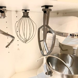 May include: A white KitchenAid stand mixer with a silver bowl and three attachments hanging from the ceiling. The attachments are a dough hook, a whisk, and a paddle.