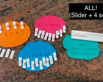 May include: Set of four alphabet and number stamp sets for baking and crafting. The sets include a slider with a variety of symbols, numbers 0-9, uppercase letters A-Z, and a set of symbols including a paw print, a flower, a heart, and a star. The stamps are white and are displayed on a variety of coloured surfaces.