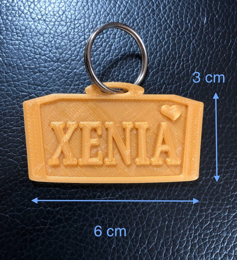 Puede incluir: Un llavero de color naranja claro con el nombre "XENIA" en letras en relieve. El llavero rectangular mide aproximadamente 6 cm de largo y 3 cm de alto, con un peque&ntilde;o detalle de coraz&oacute;n. Un llavero plateado est&aacute; adjunto en la parte superior.