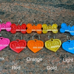 Puede incluir: Una colecci&oacute;n de etiquetas de identificaci&oacute;n para mascotas de colores, en forma de hueso y coraz&oacute;n. Las etiquetas son de color rosa, rojo, naranja, dorado y azul claro. Cada etiqueta tiene el nombre "XENIA" y un n&uacute;mero de tel&eacute;fono.