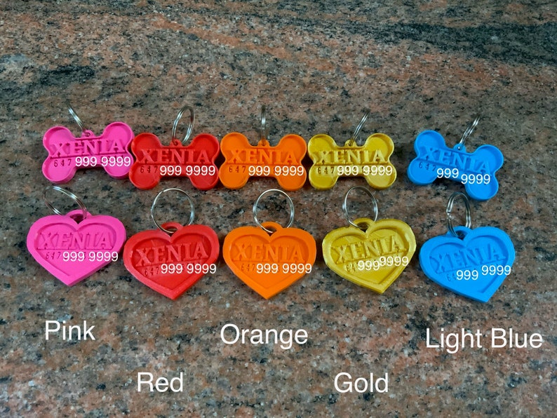 Puede incluir: Una colecci&oacute;n de etiquetas de identificaci&oacute;n para mascotas de colores, en forma de hueso y coraz&oacute;n. Las etiquetas son de color rosa, rojo, naranja, dorado y azul claro. Cada etiqueta tiene el nombre "XENIA" y un n&uacute;mero de tel&eacute;fono.