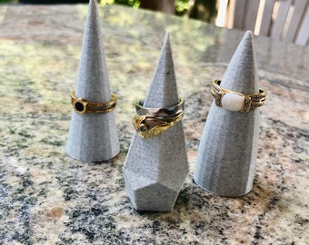 May include: Three gray cone-shaped ring holders with three different rings on top. The rings are gold and silver with different stones.