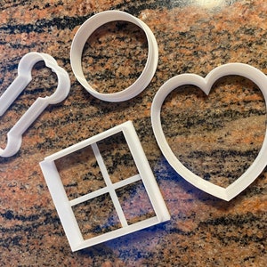 May include: Set of five white plastic cookie cutters in different shapes: a bone, a circle, a heart, and a square with four smaller squares inside.