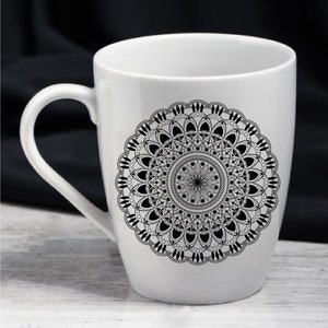 Op de afbeelding: Een witte keramische mok met een zwart-wit mandala-ontwerp. De mandala is ingewikkeld en gedetailleerd, met een cirkelvormig patroon van geometrische vormen en bloemmotieven.