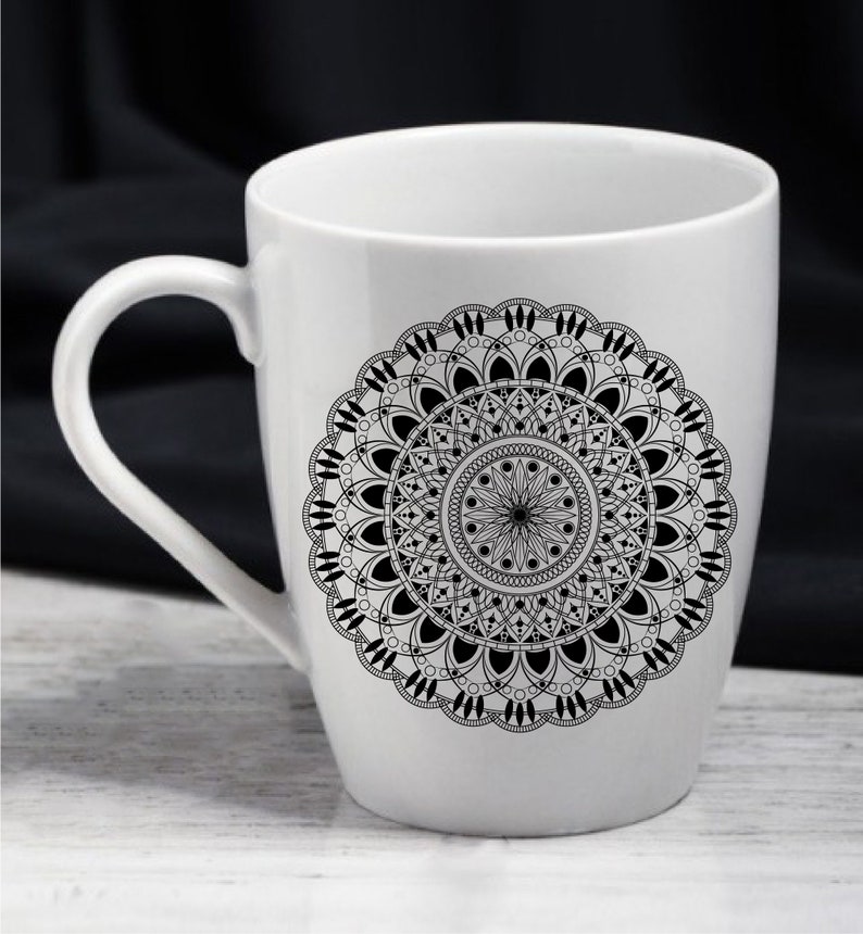 Op de afbeelding: Een witte keramische mok met een zwart-wit mandala-ontwerp. De mandala is ingewikkeld en gedetailleerd, met een cirkelvormig patroon van geometrische vormen en bloemmotieven.