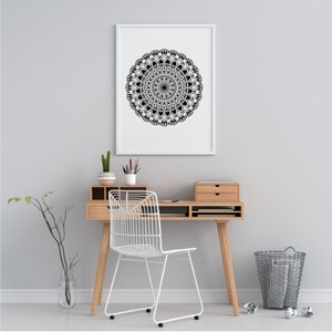 Op de afbeelding: Een zwart-wit mandala-ontwerp print in een wit frame dat aan een witte muur hangt boven een houten bureau met een witte stoel en een draad prullenbak.