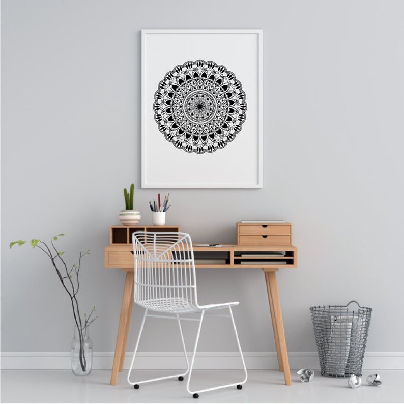 Op de afbeelding: Een zwart-wit mandala-ontwerp print in een wit frame dat aan een witte muur hangt boven een houten bureau met een witte stoel en een draad prullenbak.