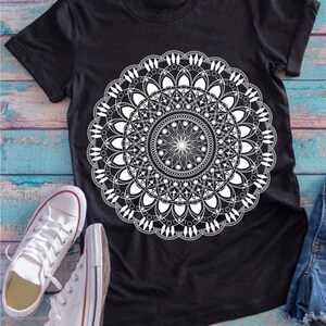 Op de afbeelding: Zwart T-shirt met een wit mandala-ontwerp. De mandala is een cirkelvormig patroon met ingewikkelde details.