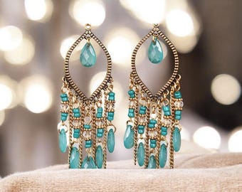 May include: A pair of ornate earrings with a bohemian design. Each earring features a teardrop-shaped turquoise stone suspended from a gold-colored frame. Below, chains of turquoise beads and clear crystals cascade downwards, creating a layered, dangling effect.