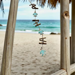 Puede incluir: Adorno colgante decorativo con tortugas marinas turquesas y pequeños trozos de madera. Las tortugas están ensartadas con hilo. El fondo muestra una playa con el mar y el cielo.