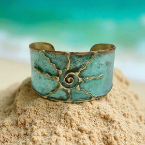 Może przedstawiać: Szeroka turkusowa bransoletka z motywem słońca. Bransoletka ma obramowanie w kolorze mosiądzu i złote akcenty. Manka leży na piasku z rozmytym tłem oceanu.