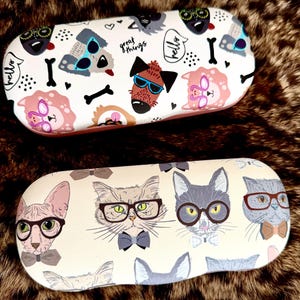 Puede incluir: Dos estuches ovalados rígidos para gafas. El estuche superior es blanco con un patrón colorido de perros con gafas de sol, huesos y el texto "great things" y "hello". El estuche inferior es crema con un patrón de gatos con gafas y pajaritas.