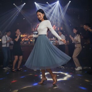 May include: A woman in a white top and a blue tulle skirt dances in a club setting. The skirt is a flowing, knee-length style. She wears heeled sandals. The background features a bar and other people enjoying the atmosphere.