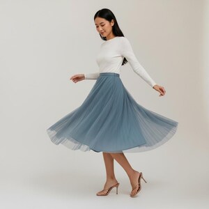 May include: A woman in a white long-sleeved top and a flowing, dusty blue tulle skirt. The skirt is pleated and falls to mid-calf length. She is wearing gold-coloured high-heeled sandals.