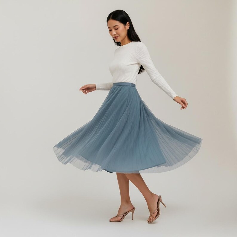 May include: A woman in a white long-sleeved top and a flowing, dusty blue tulle skirt. The skirt is pleated and falls to mid-calf length. She is wearing gold-coloured high-heeled sandals.