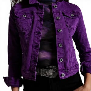 May include: A striking purple denim jacket with a traditional button-up design. The jacket has two chest pockets and buttoned cuffs. The model is wearing a black top and a belt with a silver buckle.
