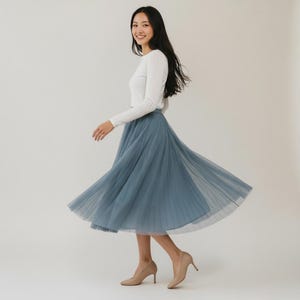 May include: A woman in a white long-sleeved top and a flowing, dusty blue tulle skirt. She is also wearing beige high heels. The skirt is knee-length and appears to be made of multiple layers of soft fabric.