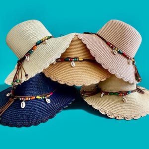 Puede incluir: Una colección de cinco sombreros de paja trenzada en azul marino, crema y beige. Cada sombrero presenta un borde festoneado y una banda de cordón de cuero marrón decorada con cuentas coloridas y pequeñas conchas blancas, con extremos colgantes.