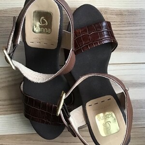 May include: A pair of brown leather sandals with a crocodile-embossed strap and a gold buckle. The sandals have a cork wedge heel and are labeled "Kanna" with "Made in Spain".