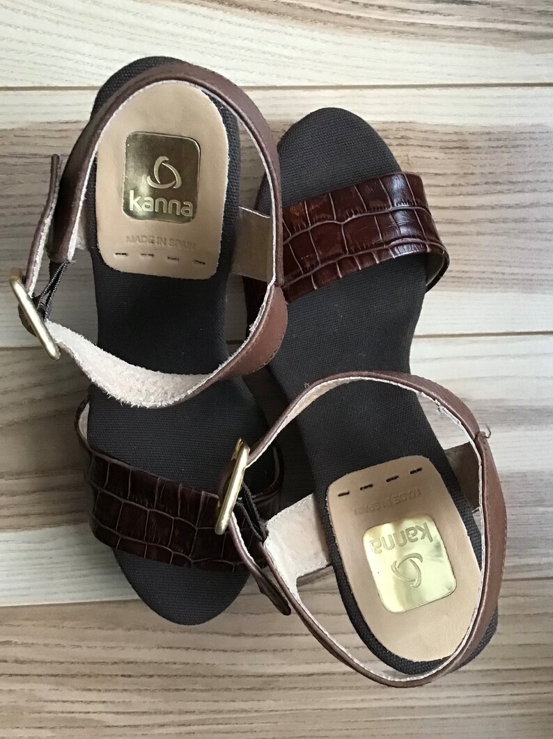 May include: A pair of brown leather sandals with a crocodile-embossed strap and a gold buckle. The sandals have a cork wedge heel and are labeled "Kanna" with "Made in Spain".