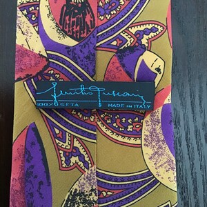 May include: A silk tie with a paisley pattern in shades of purple, red, and gold. The tie has a black label with the text "Emilio Pucci" and "100% Seta" and "Made in Italy".