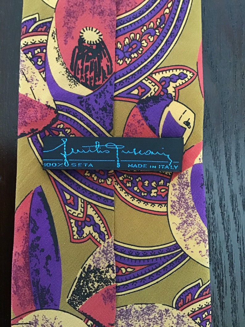 May include: A silk tie with a paisley pattern in shades of purple, red, and gold. The tie has a black label with the text "Emilio Pucci" and "100% Seta" and "Made in Italy".