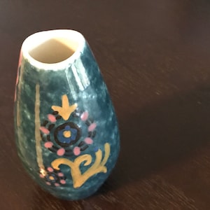 May include: A small, teal-colored ceramic vase with a white rim. The vase has a floral design in pink, yellow, blue, and black.