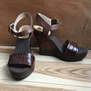 May include: A pair of brown wedge sandals with an ankle strap and a crocodile-embossed design. The sandals have a woven wedge heel and a brown canvas sole.