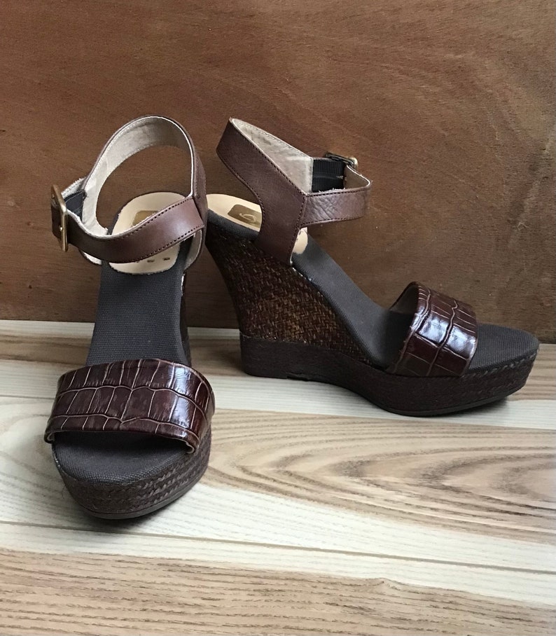 May include: A pair of brown wedge sandals with an ankle strap and a crocodile-embossed design. The sandals have a woven wedge heel and a brown canvas sole.