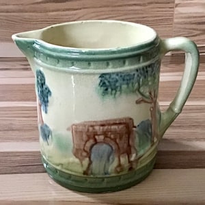 May include: A vintage ceramic pitcher with a green and yellow glaze. The pitcher features a hand-painted scene of a wagon pulled by two oxen. The scene is surrounded by trees and a green border.