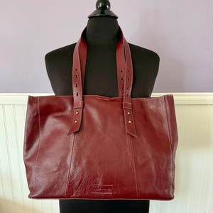 May include: A burgundy leather tote bag with two top handles and a rectangular shape. The bag has white stitching and gold-colored rivets. The bag is displayed on a black mannequin.