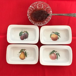 May include: Four white ceramic rectangular dishes with fruit designs. Each dish has a different fruit: plums, pears, apples, and peaches. The dishes are on a red surface.